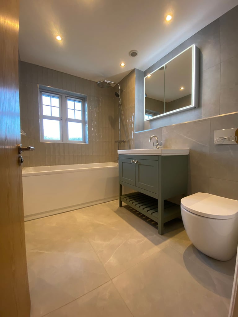 Modern bathroom with green vanity, white toilet, bathtub, illuminated mirror, and neutral tile flooring
