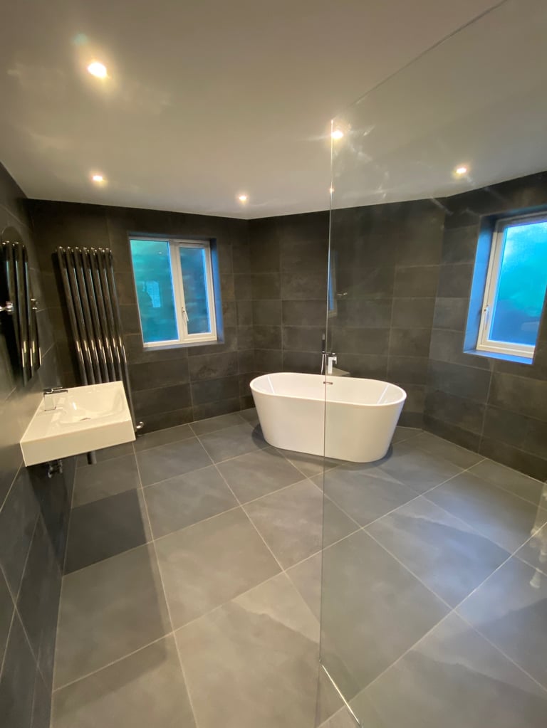 Modern luxury bathroom with freestanding white bathtub, large frameless glass shower enclosure, dark tile walls, and recessed ceiling lights