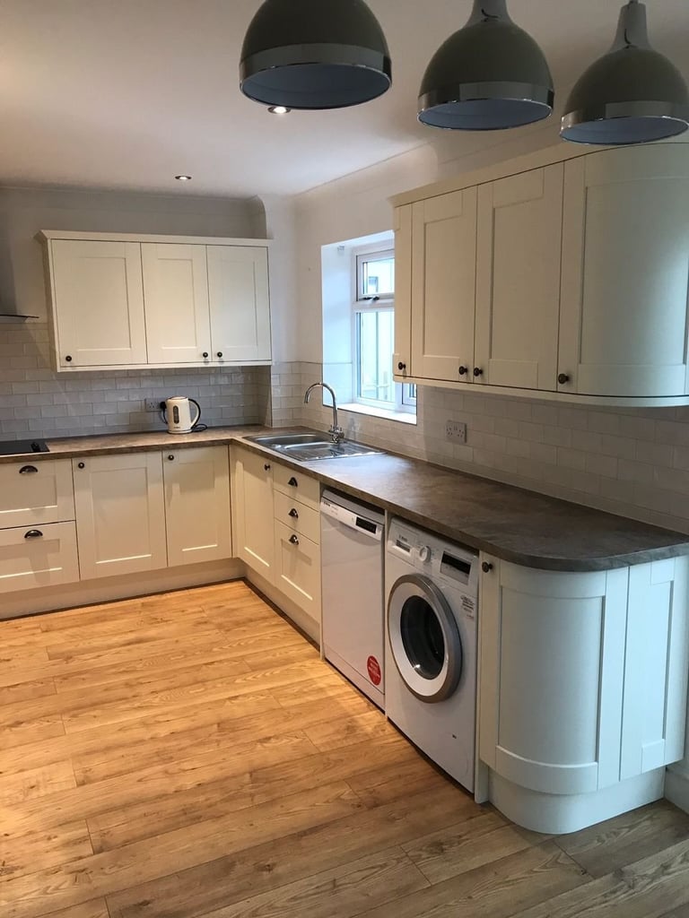 Modern kitchen with cream cabinets, dark countertops, black pendant lights, wooden flooring, and white washing machine