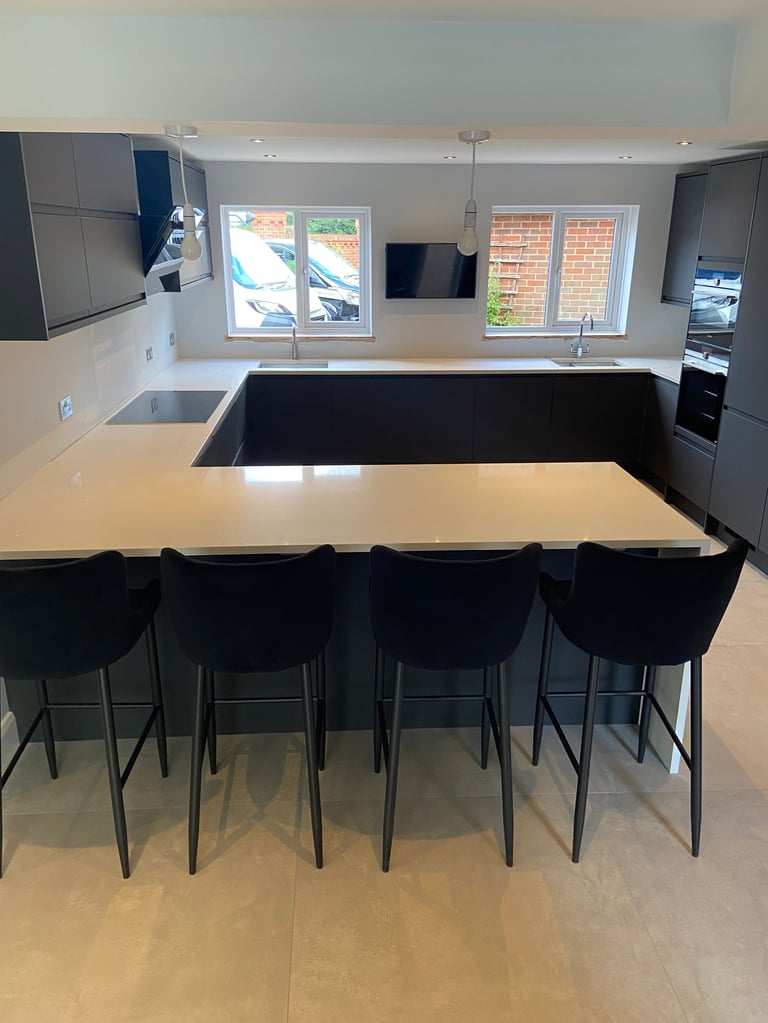 Modern kitchen with large island, black bar stools, white countertop, gray cabinetry, and windows overlooking brick exterior